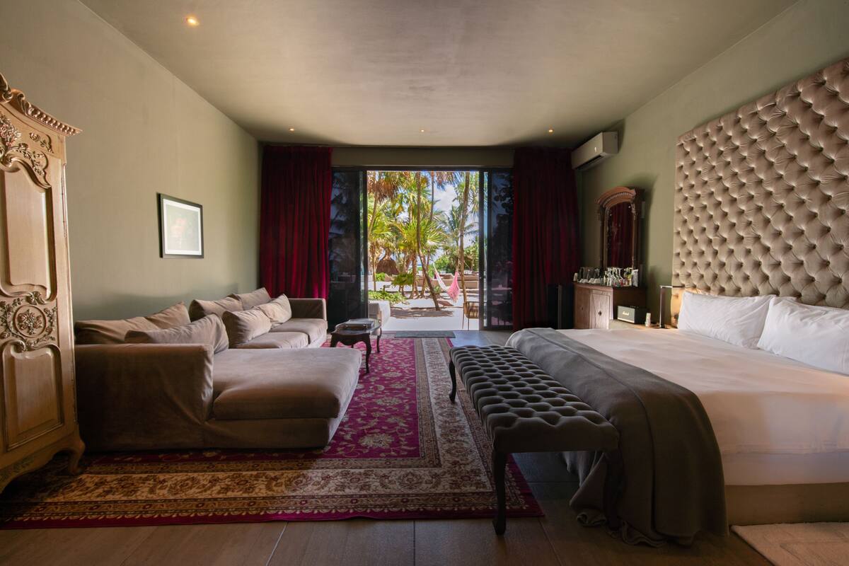 Une chambre spacieuse avec un grand lit, un canapé et un banc. La fenêtre offre une vue sur les palmiers.