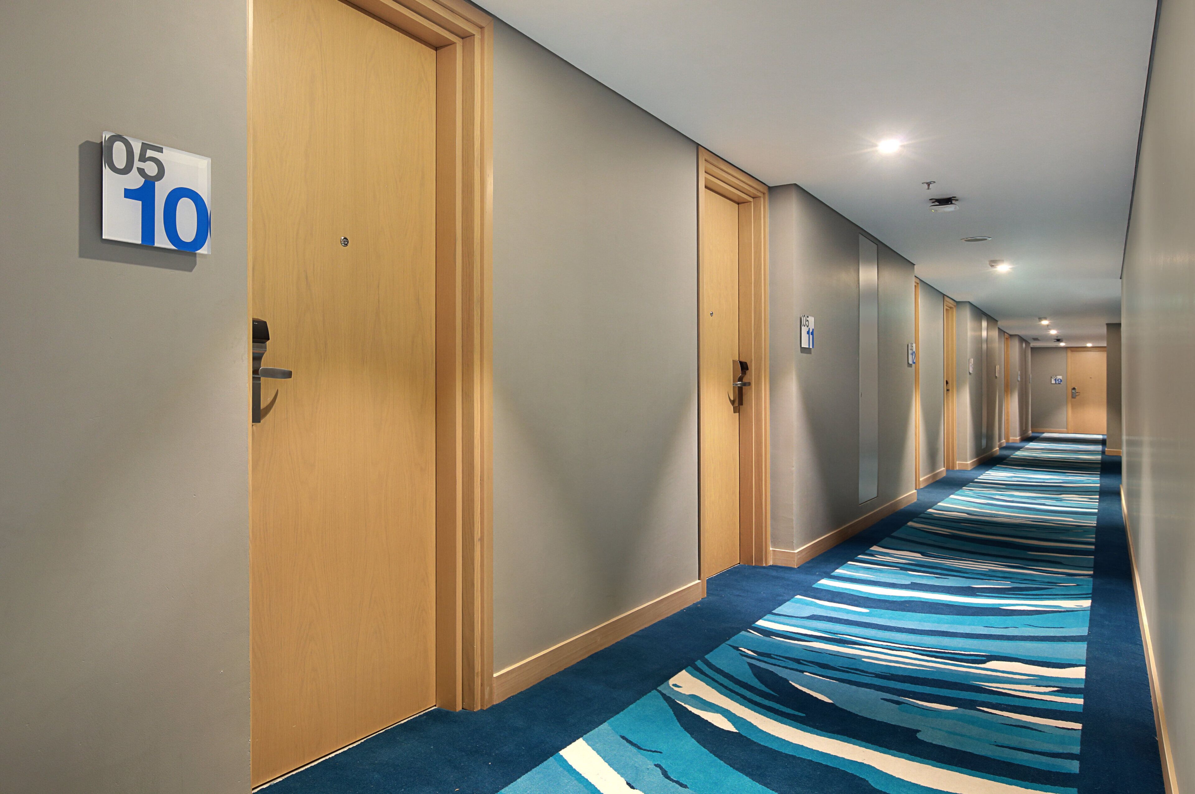 Corredor de hotel com portas numeradas de 05 a 10, com carpete estampado em azul e branco.