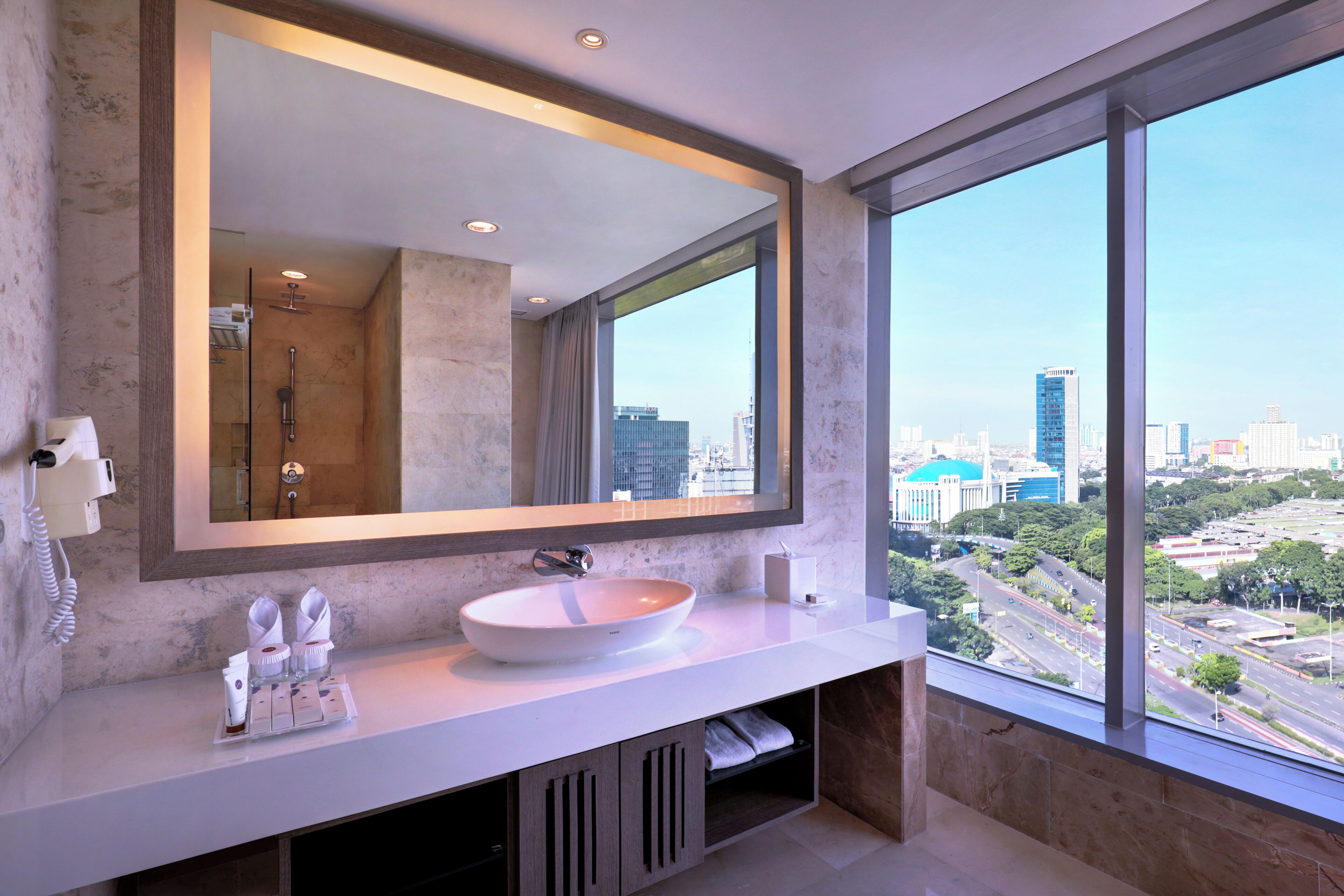 Un baño moderno con un espejo grande, un lavabo blanco y vistas a un paisaje urbano a través de un ventanal amplio.