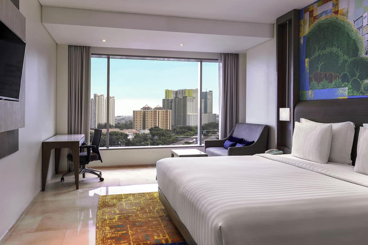A hotel room with a large bed, a desk, a chair, and a view of city buildings.