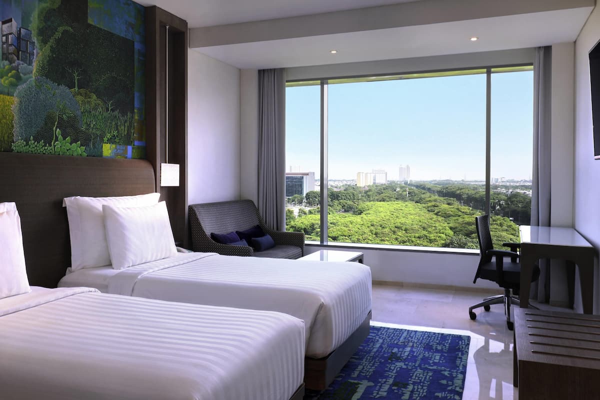 A hotel room with two beds, a desk, a chair, and a large window offering a city view.