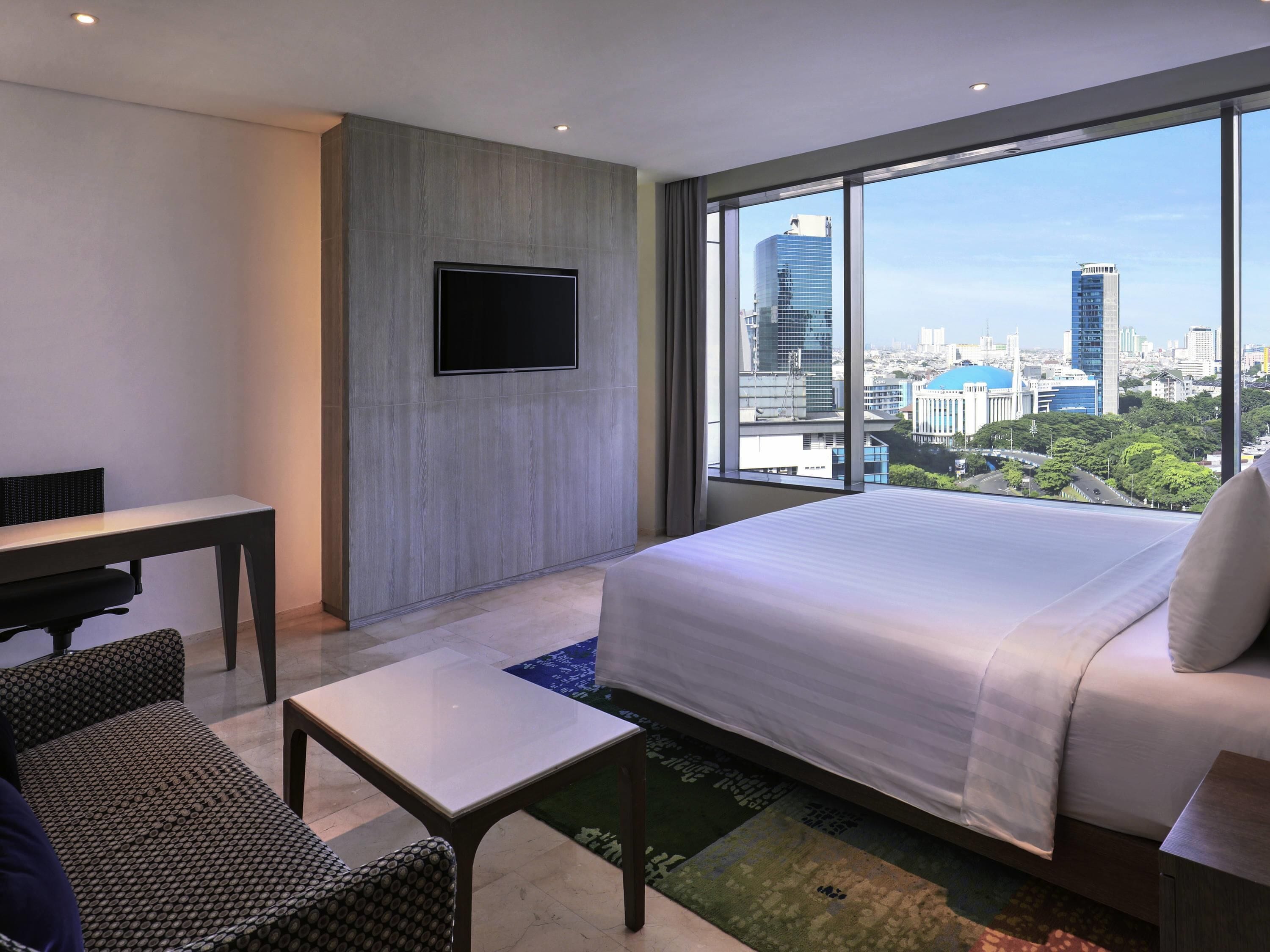 Una camera d'albergo con un letto grande, una scrivania, una sedia, una TV a parete e vista sulla città attraverso ampie finestre.
