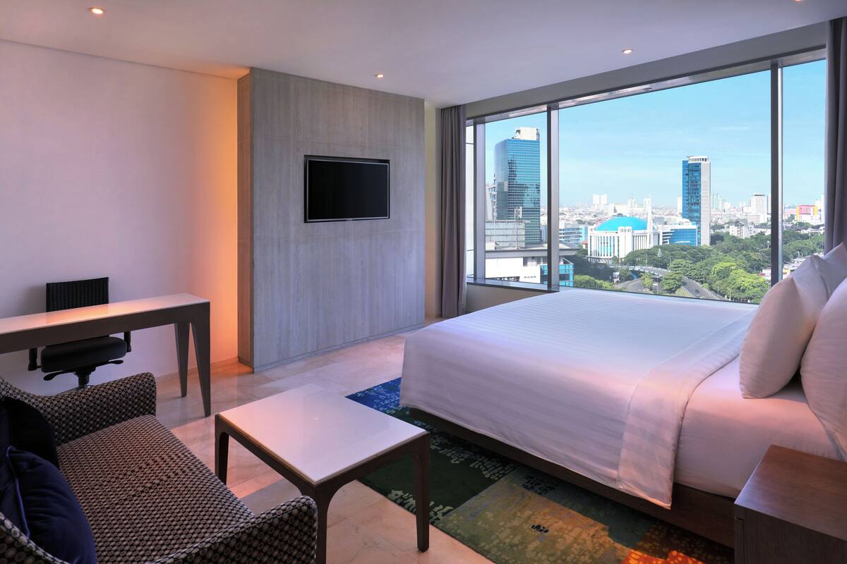 A hotel room with a large bed, a desk, a chair, a TV, and a city view.