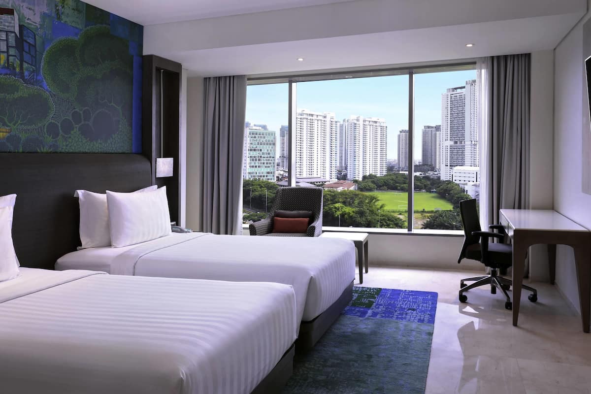 A hotel room with two beds, a desk, and a large window offering a city view.