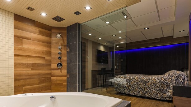 A modern bathroom with a bathtub, wooden wall panels, a glass shower enclosure, and a bed with a patterned cover. - Hôtel Le Chablis (Montreal)