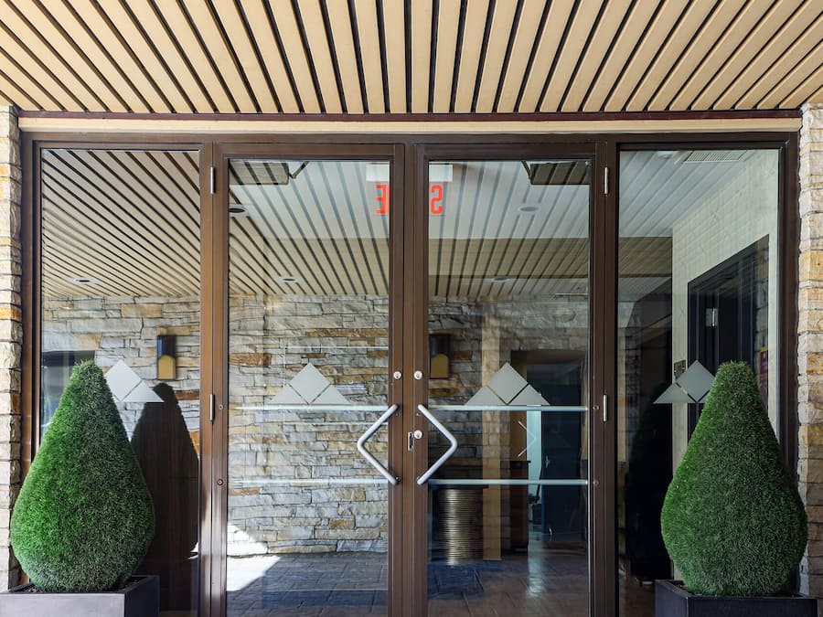 Entrada de hotel com paredes de pedra, teto de madeira e porta de vidro com desenhos geométricos.