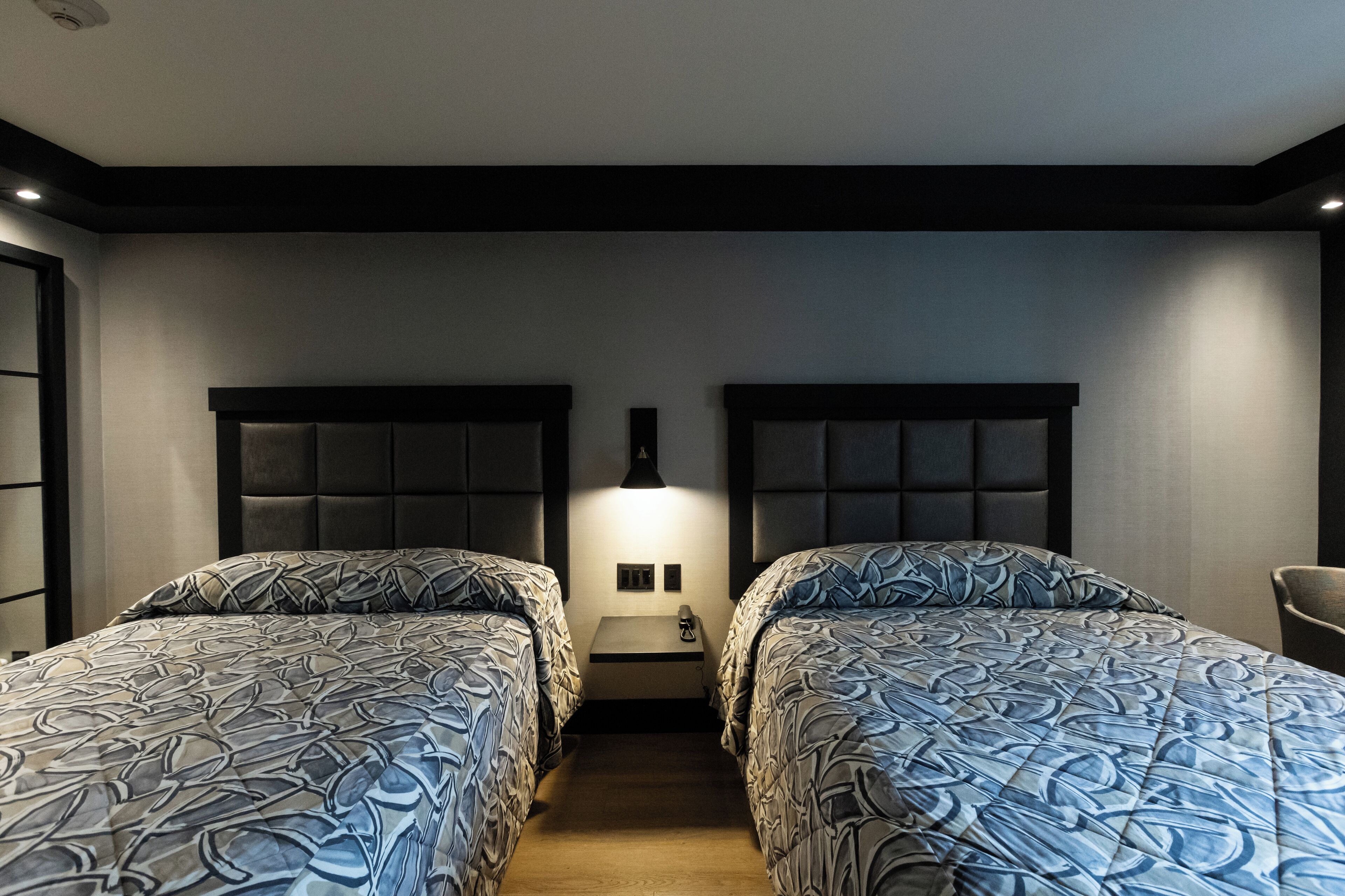 A hotel room with two beds, each with a headboard and a patterned bedspread. There is a small bedside table between the beds with a lamp and a phone.