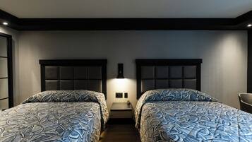A hotel room with two beds, each with a headboard and a patterned bedspread. There is a small bedside table between the beds with a lamp and a phone.