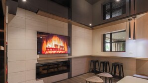 A modern kitchen with a flat-screen TV displaying a fireplace, wooden bar stools, and a window with a view. - Hôtel Le Chablis (Montreal)