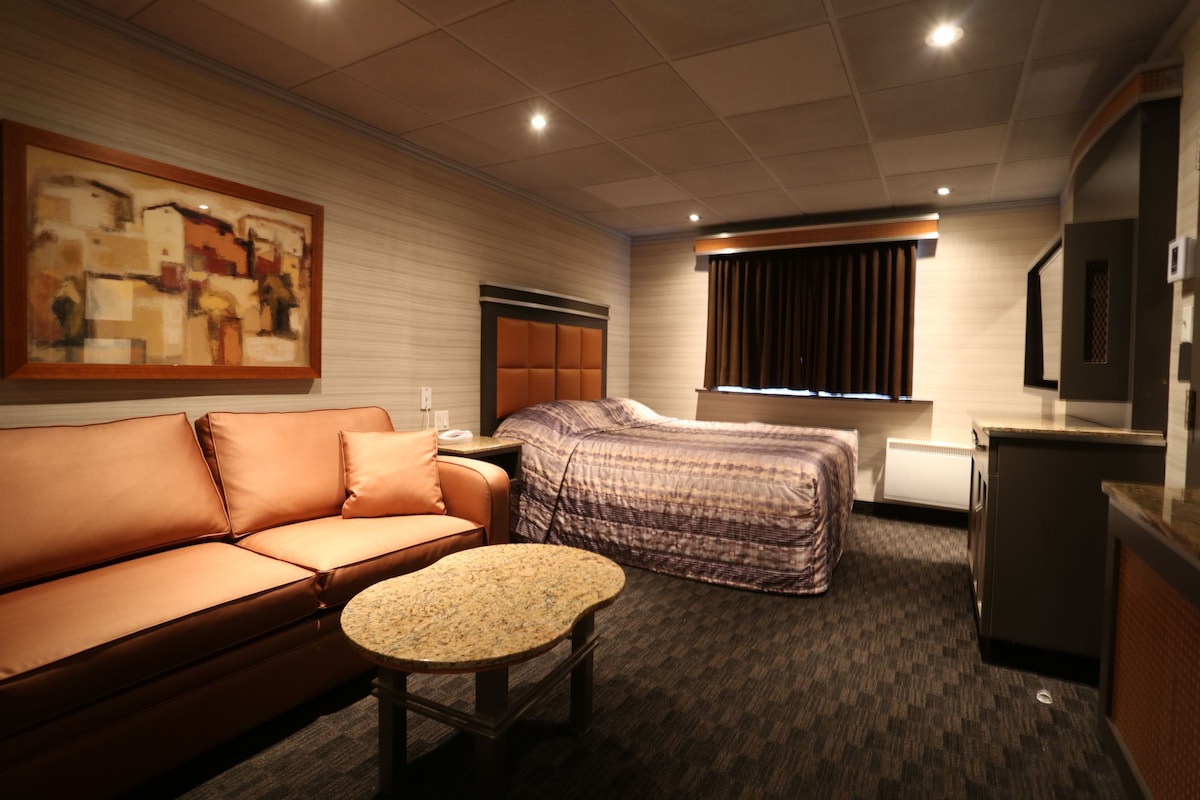 A hotel room with a sofa, a bed, a small table, a painting on the wall, a window with curtains, and a television.