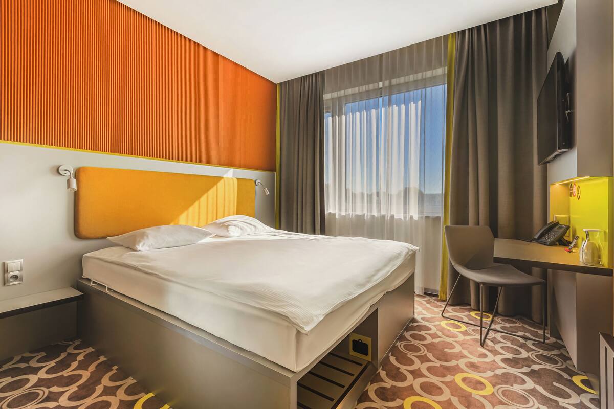 Habitación de hotel con cama, escritorio, silla, televisor y ventana con cortinas.