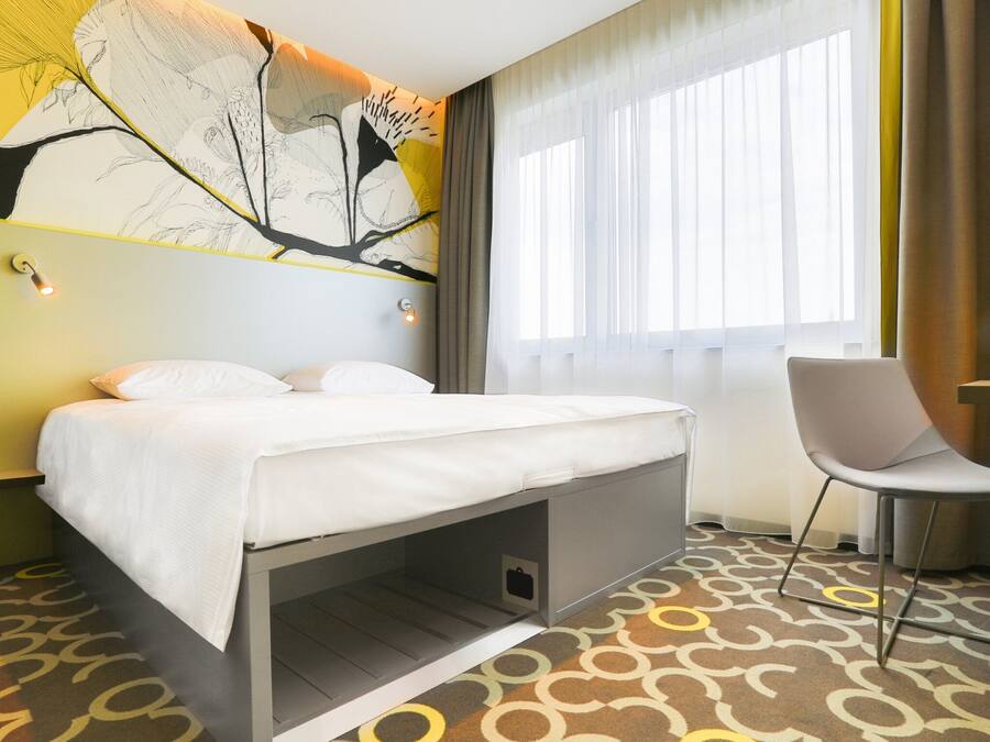 A hotel room with a bed, a chair, a large window with sheer curtains, and a wall mural.