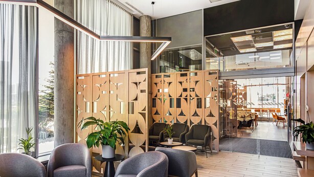 A modern lobby with grey armchairs, a wooden partition with geometric patterns, and a view of the outdoor seating area. - Qubus Hotel Żory (Zory)