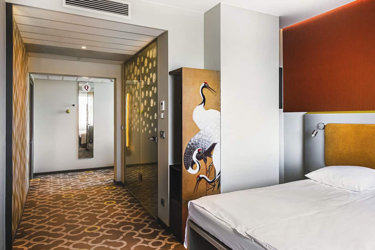 Una habitación de hotel con alfombra estampada, cama con ropa de cama blanca, un cuadro de un pájaro colgado en la pared y una puerta que da a otra habitación.