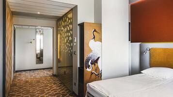 A hotel room with a patterned carpet, a bed with white linens, a wall-mounted artwork of a bird, and a door leading to another room.