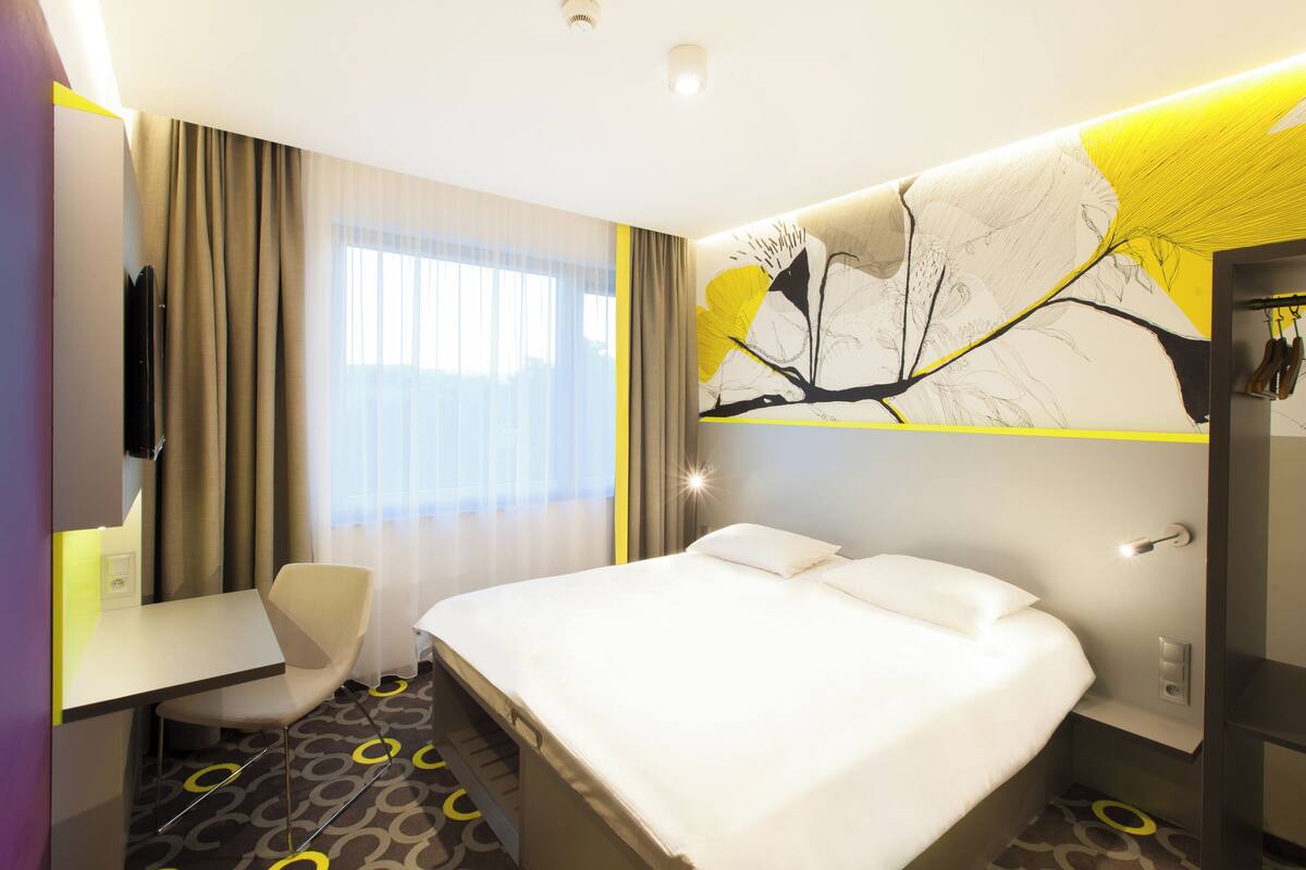 Una habitación de hotel con cama, escritorio, silla y un amplio ventanal con cortinas.