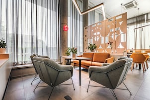 A modern hotel lobby with grey upholstered chairs, an orange sofa, and a wooden partition with geometric cutouts.