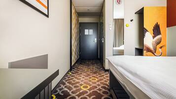 A hotel room with a bed, a bathroom with a mirror, a door, and a hallway with patterned flooring.