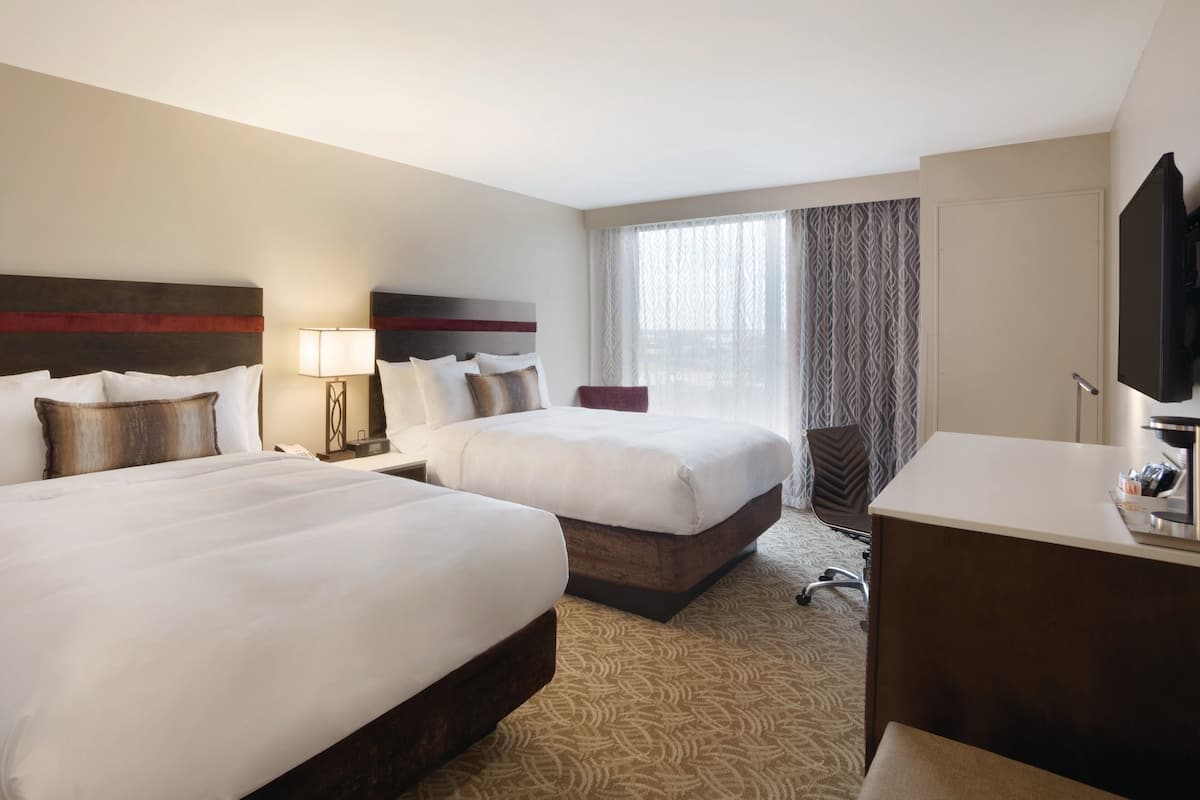 A hotel room with two beds, a desk, a chair, a TV, and a large window with curtains.
