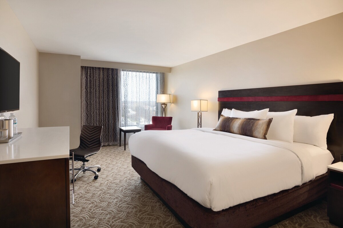 Een hotelkamer met een groot bed, een bureau met een flatscreen televisie, een stoel, een raam met gordijnen en twee bedlampjes.