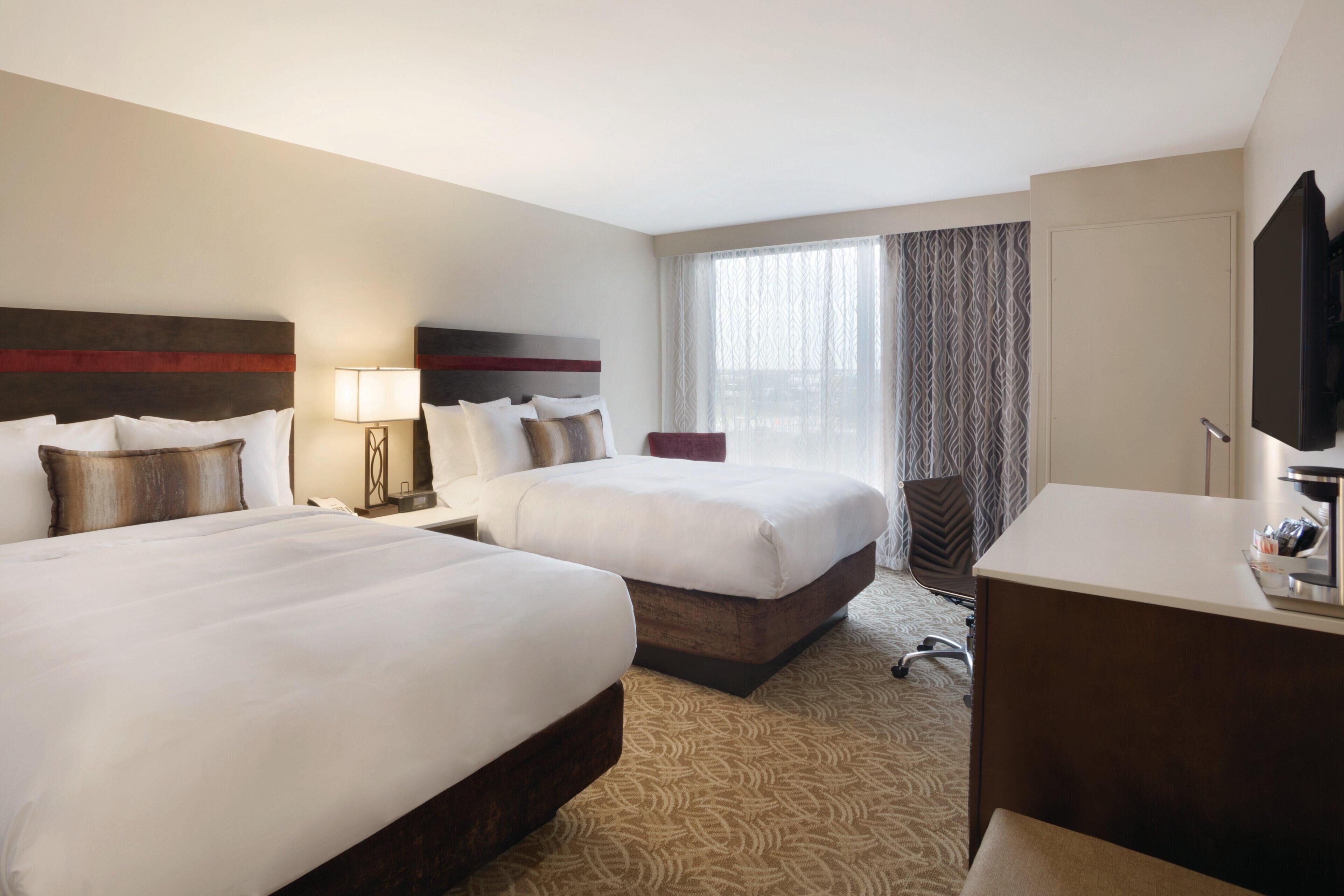 A hotel room with two beds, a desk, a chair, a TV, and a window with curtains.