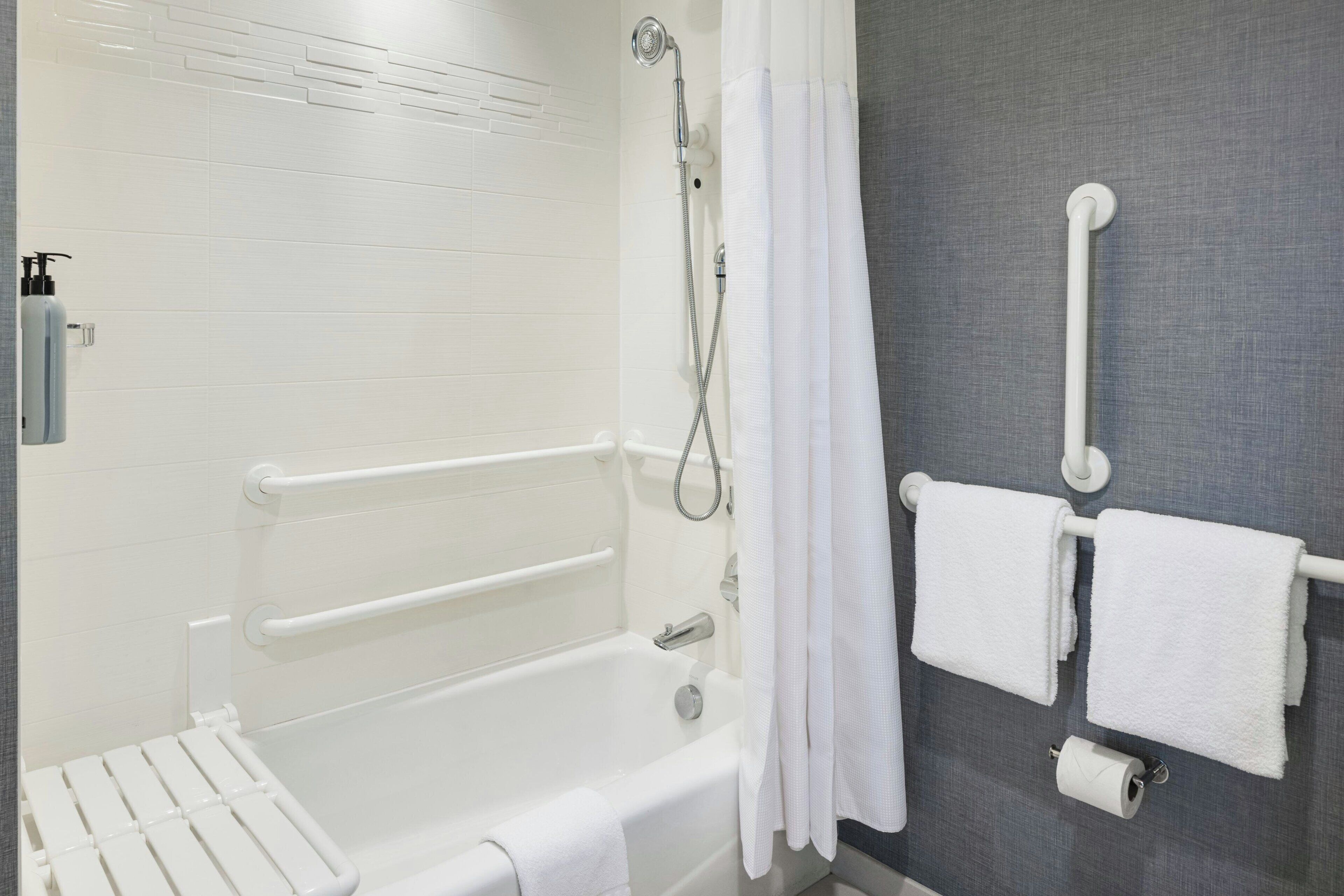 A bathroom with a white bathtub, grab bars, a showerhead, a towel rack, and a toilet paper holder.
