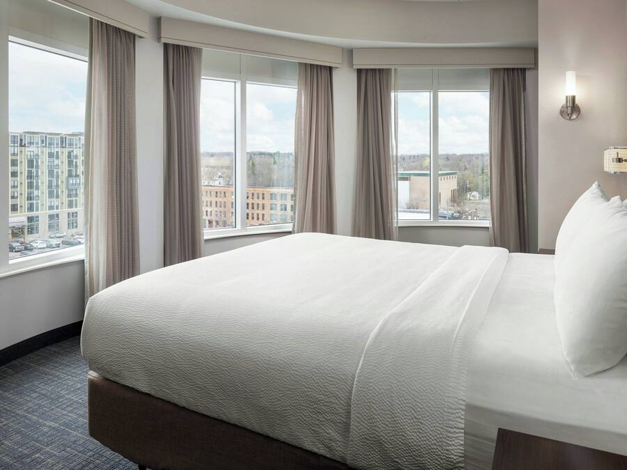 A hotel room with a large bed, multiple windows, and a city view.