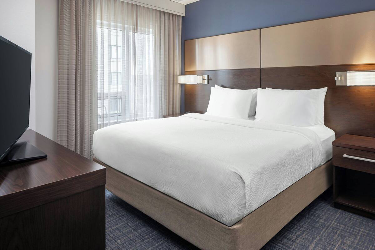 A hotel room with a large bed, a bedside table with a lamp, a TV on a desk, and a window with curtains.