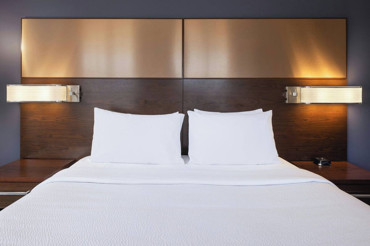 A neatly made bed with white linens, a wooden headboard, and two wall-mounted lamps on either side.