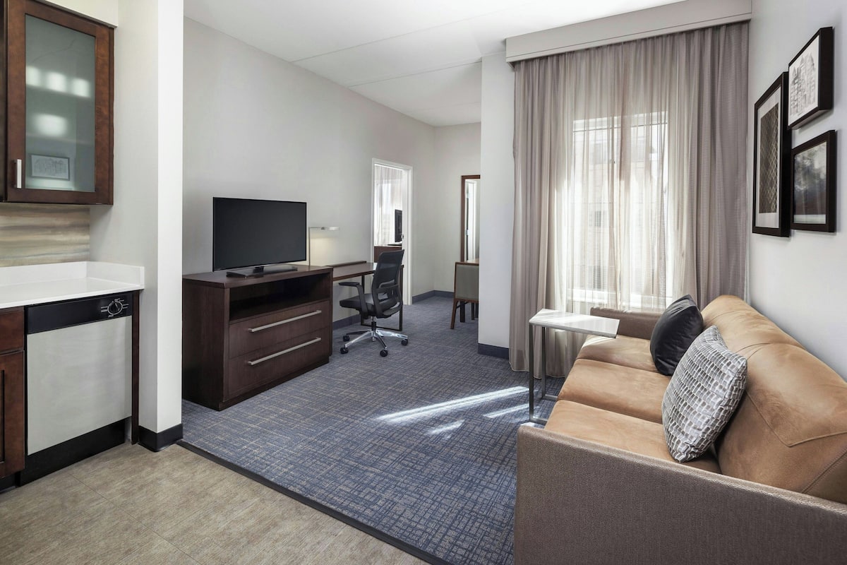 A hotel room with a kitchenette, a flat-screen TV, a sofa, a desk, and a large window with curtains.