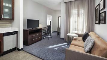 A hotel room with a kitchenette, a flat-screen TV, a sofa, a desk, and a large window with curtains.