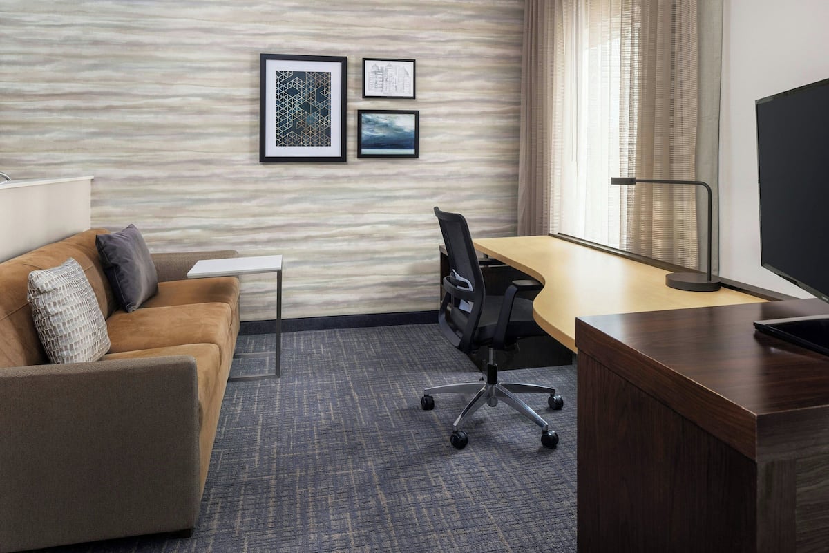 A hotel room with a wooden desk, a black office chair, a brown sofa with cushions, a small white table, and a framed picture on the wall.