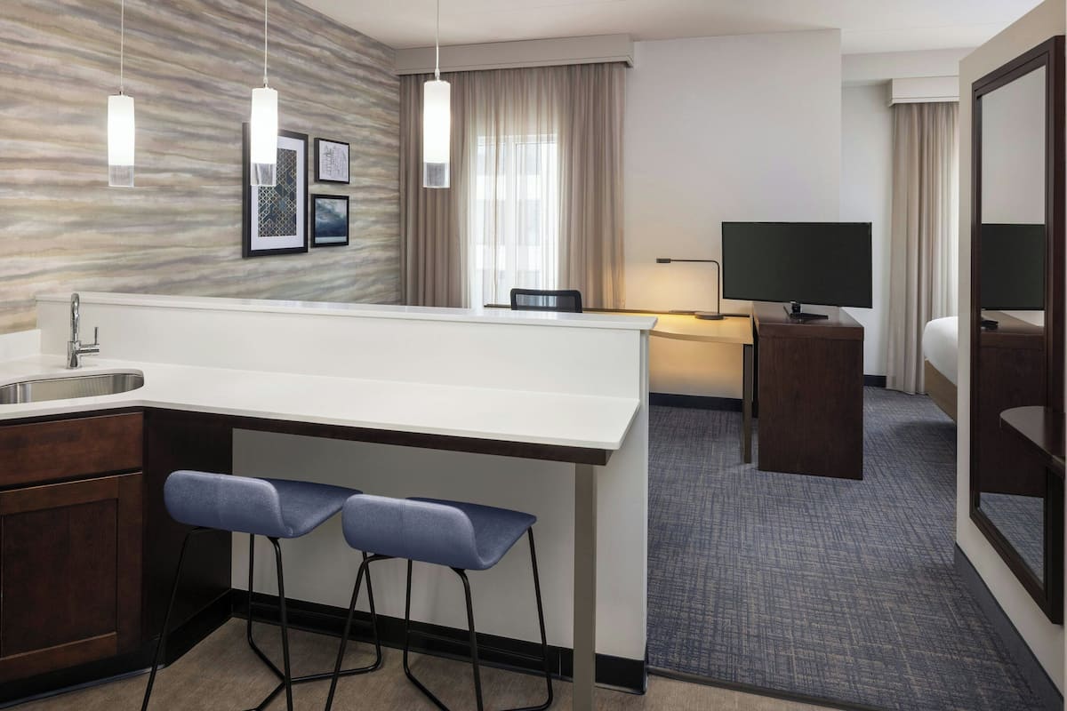 A modern hotel room with a kitchen area, a dining table, two bar stools, a flat-screen TV, and a large mirror.