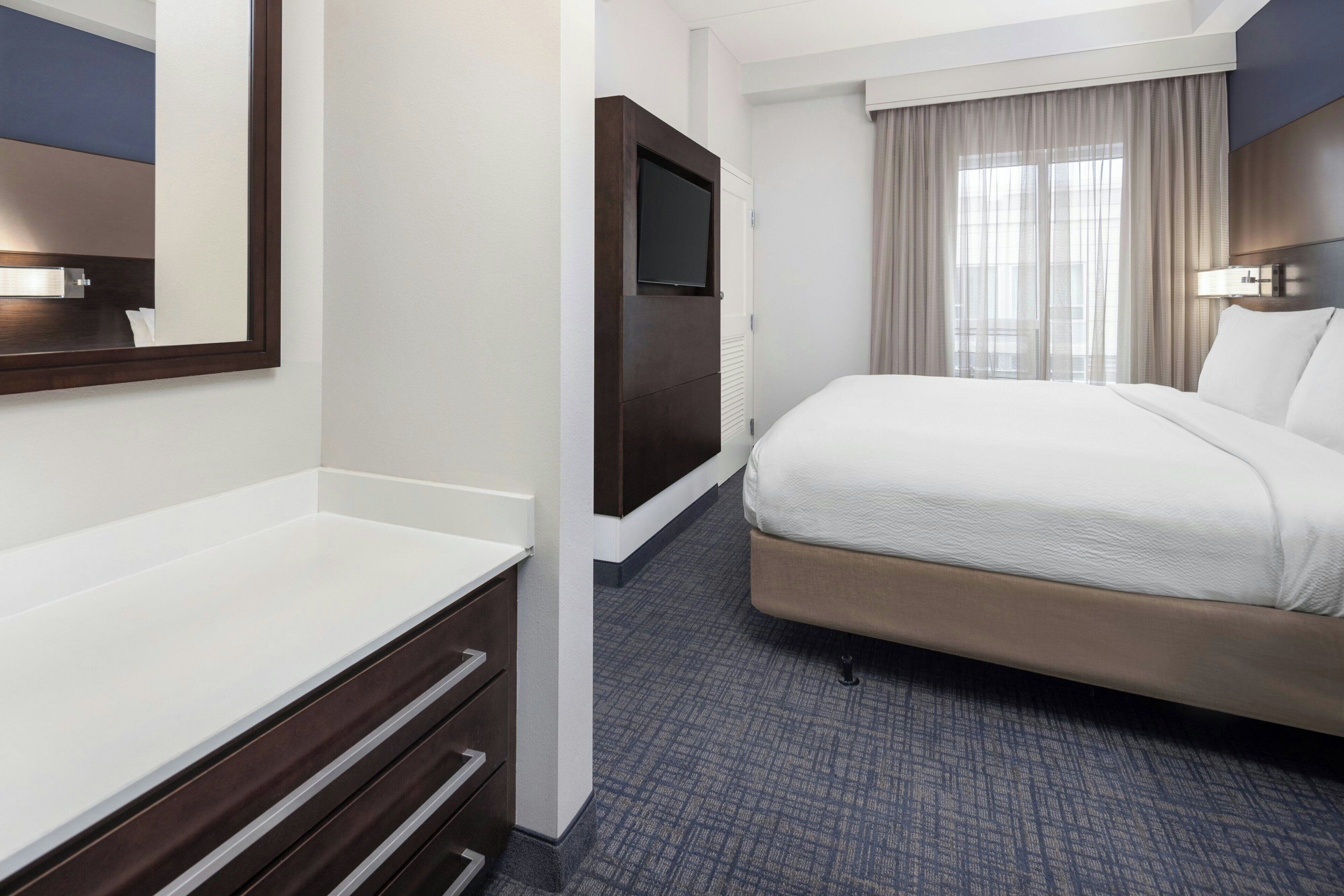 A hotel room with a bed, a TV mounted on a wall, a desk with drawers, and a window with curtains.