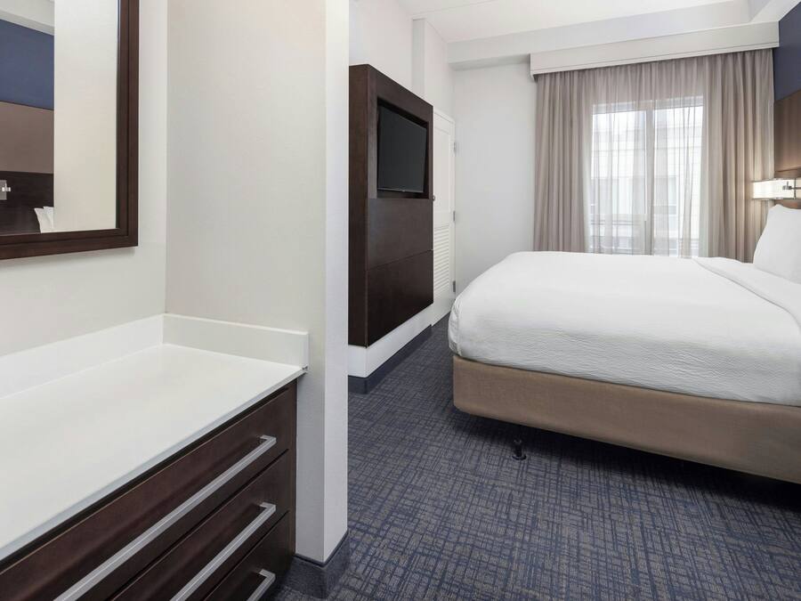 A hotel room with a bed, a TV mounted on a wall, a desk with drawers, and a window with curtains.