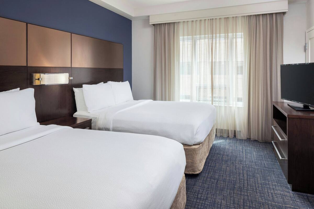 A hotel room with two beds, a TV, a desk, and a large window with curtains.