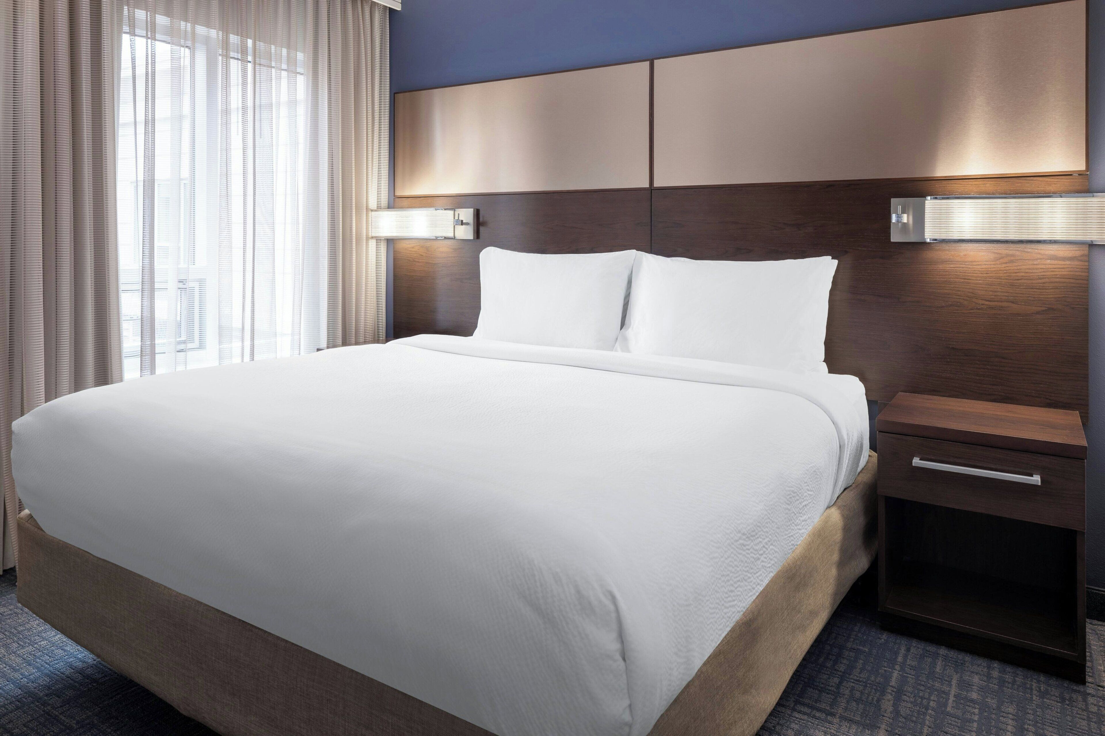 A hotel room with a large bed, a wooden headboard, a nightstand with a drawer, and a window with sheer curtains.