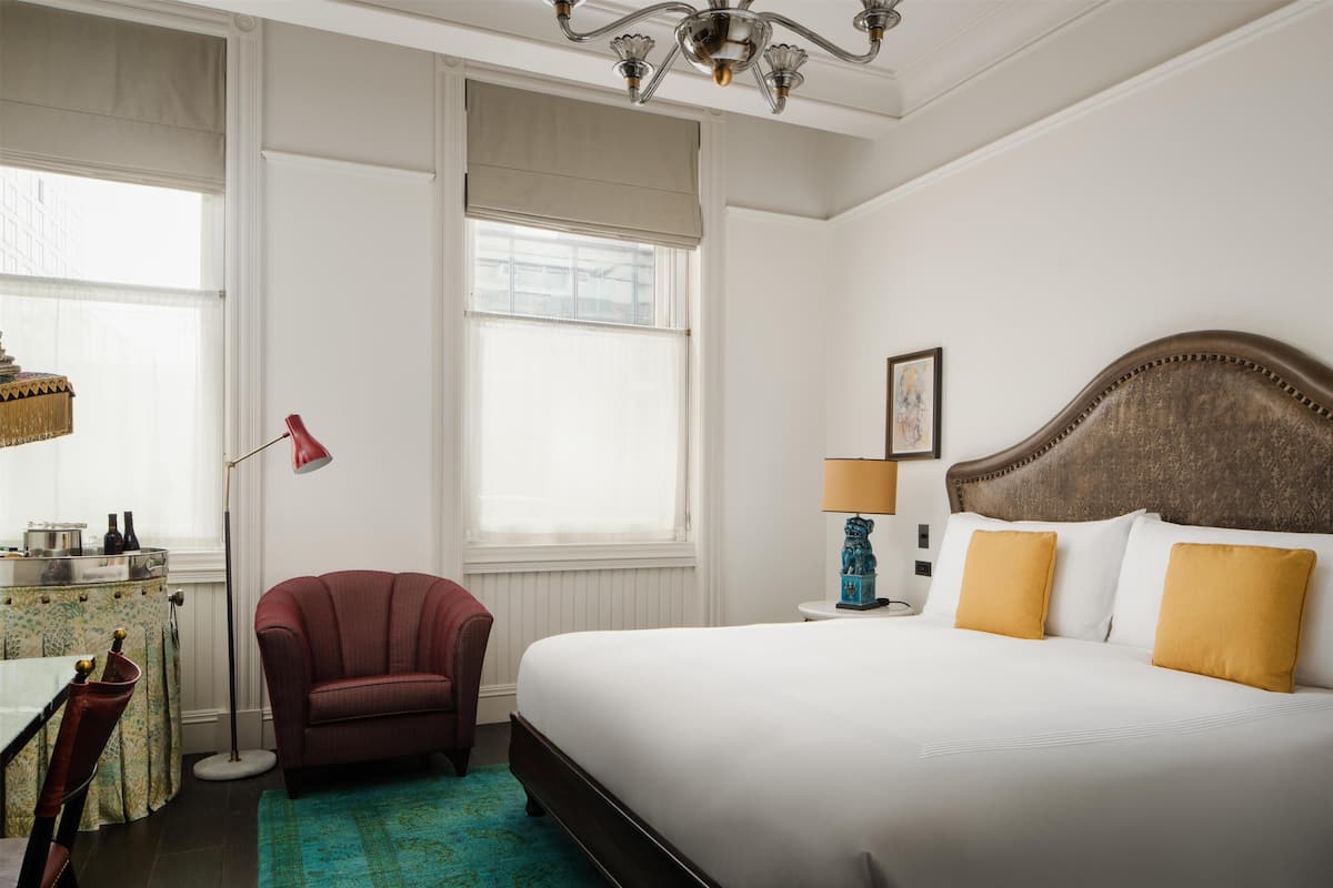 Quarto de hotel com cama, cadeira, luminária, mesa de cabeceira e janela.