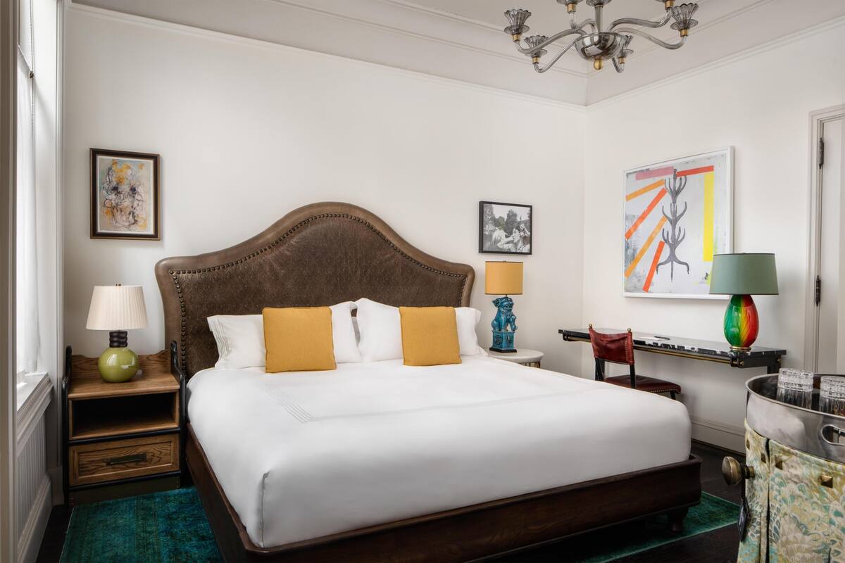 Quarto de hotel com uma cama grande, mesa de cabeceira com abajur, uma cadeira, escrivaninha com luminária e obras de arte nas paredes.