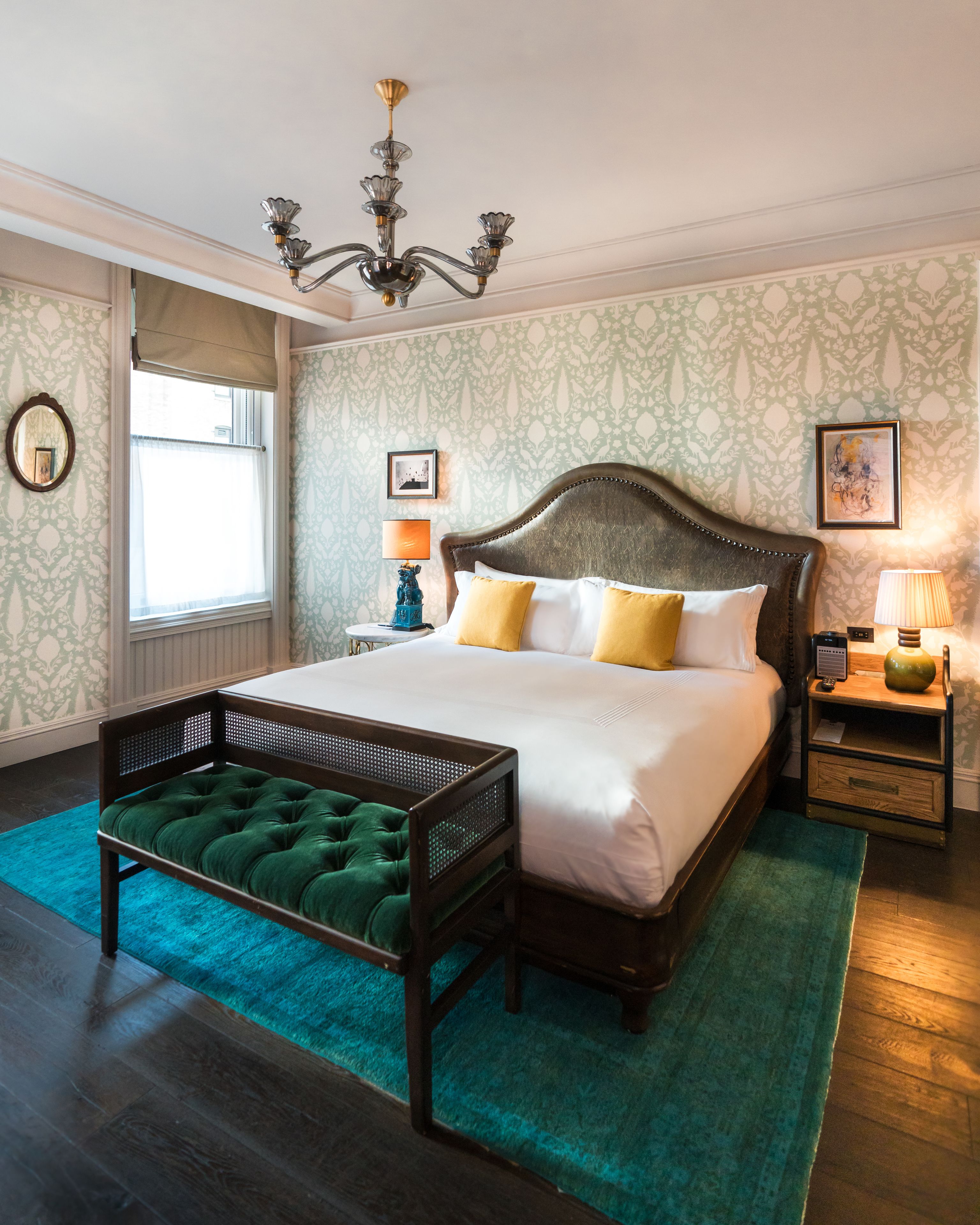 A spacious bedroom with a large bed, a wooden headboard, a green upholstered bench, a chandelier, and patterned wallpaper.