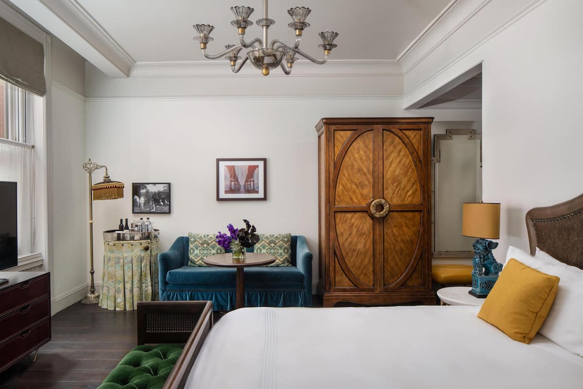 Quarto de hotel com uma cama, um sofá azul, um guarda-roupa de madeira, uma mesa redonda com um vaso e um candelabro.