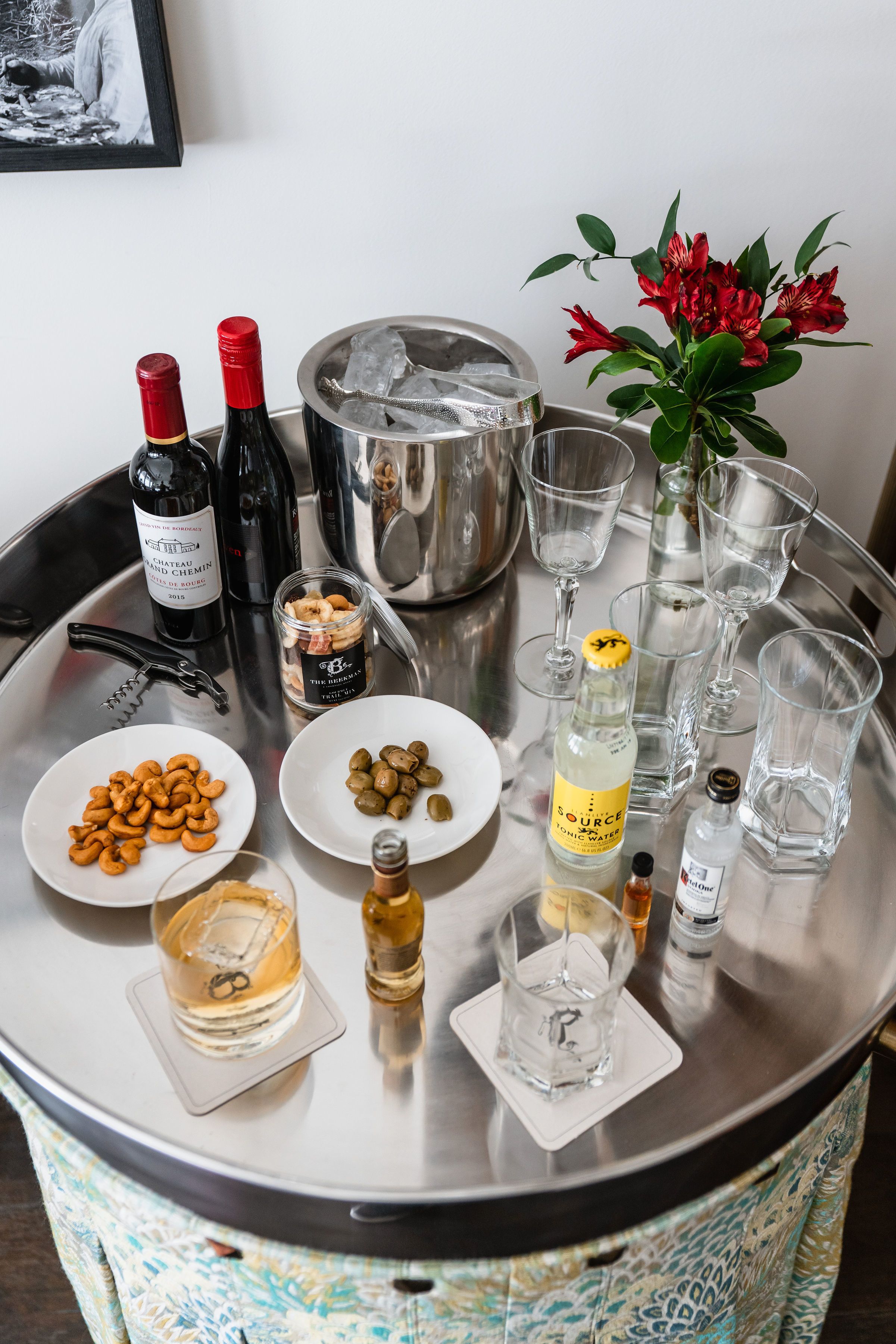 A round table with a variety of items including wine bottles, glasses, a jar of nuts, a bottle of tonic water, and a vase with red flowers.