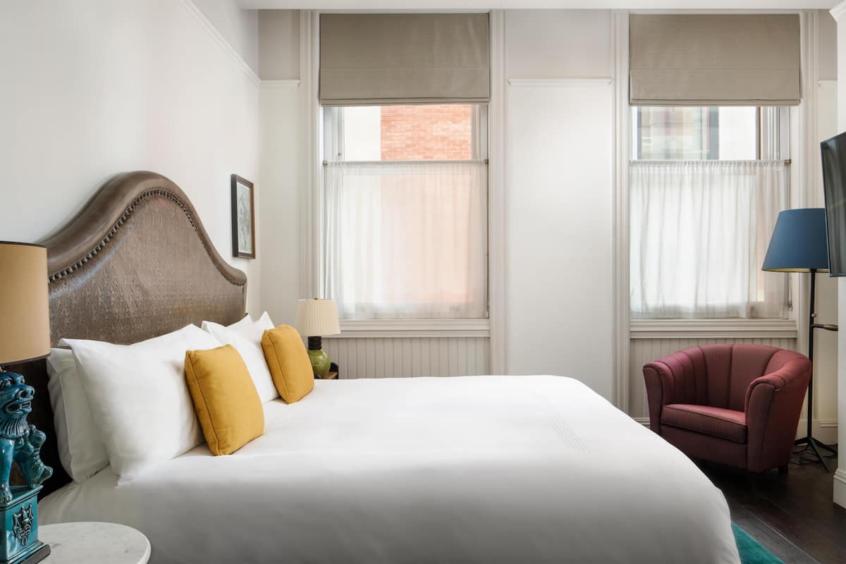 Quarto de hotel com uma cama grande, duas janelas com cortinas, uma poltrona vermelha e um abajur.
