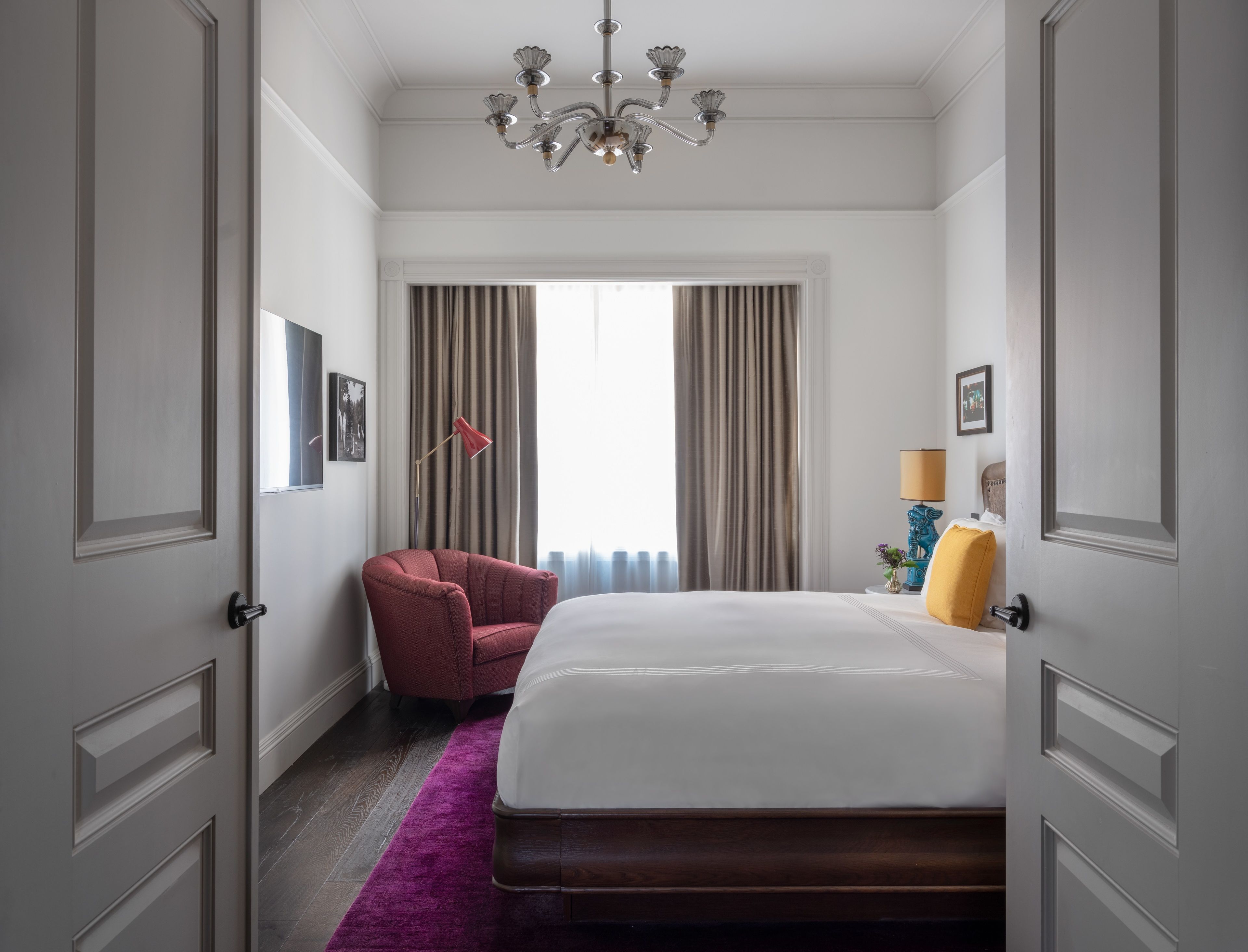 Une chambre d’hôtel avec un grand lit, un fauteuil rouge, un lustre et une vue sur l’extérieur par la fenêtre.