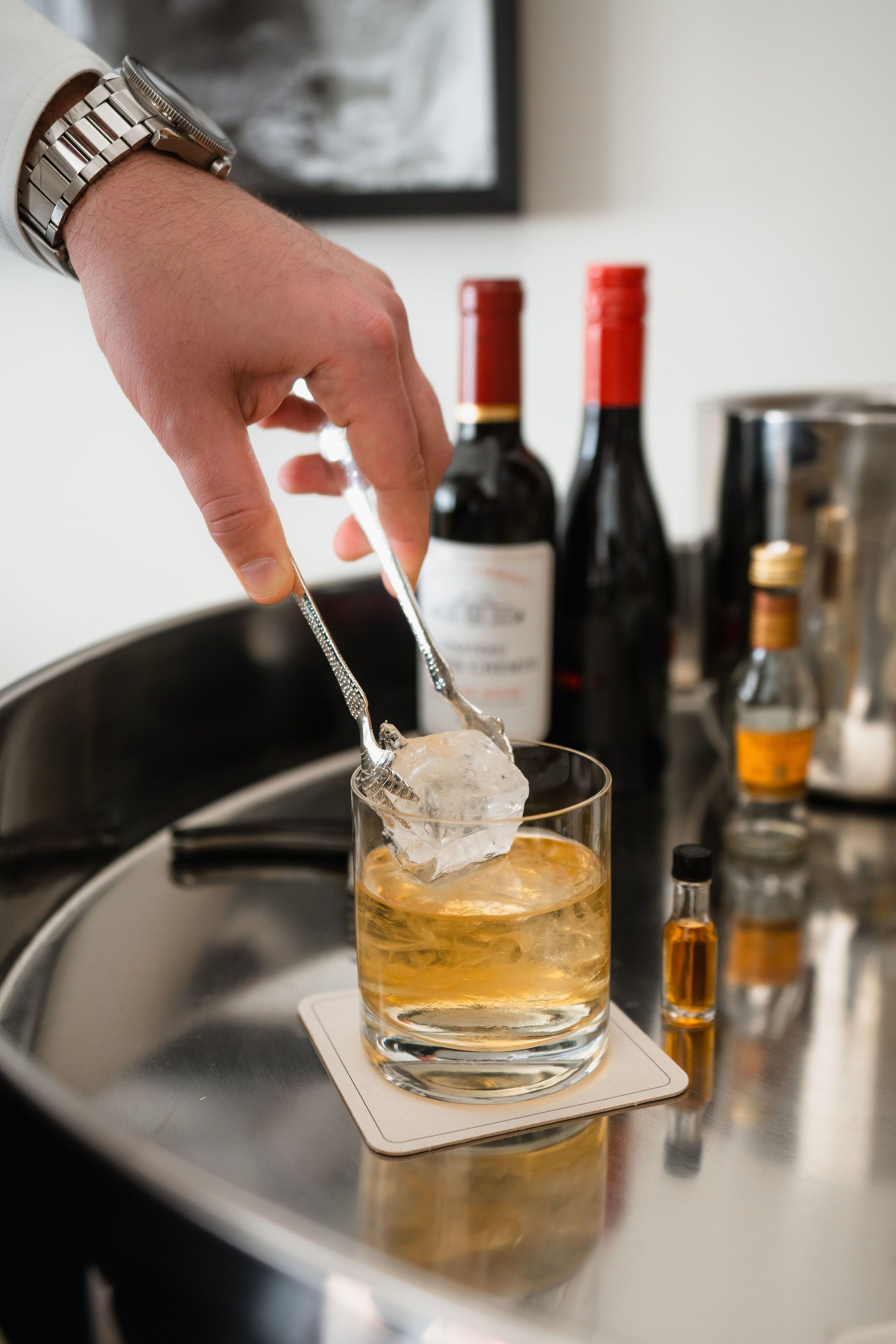 A hand holding a spoon with ice cubes over a glass on a coaster.
