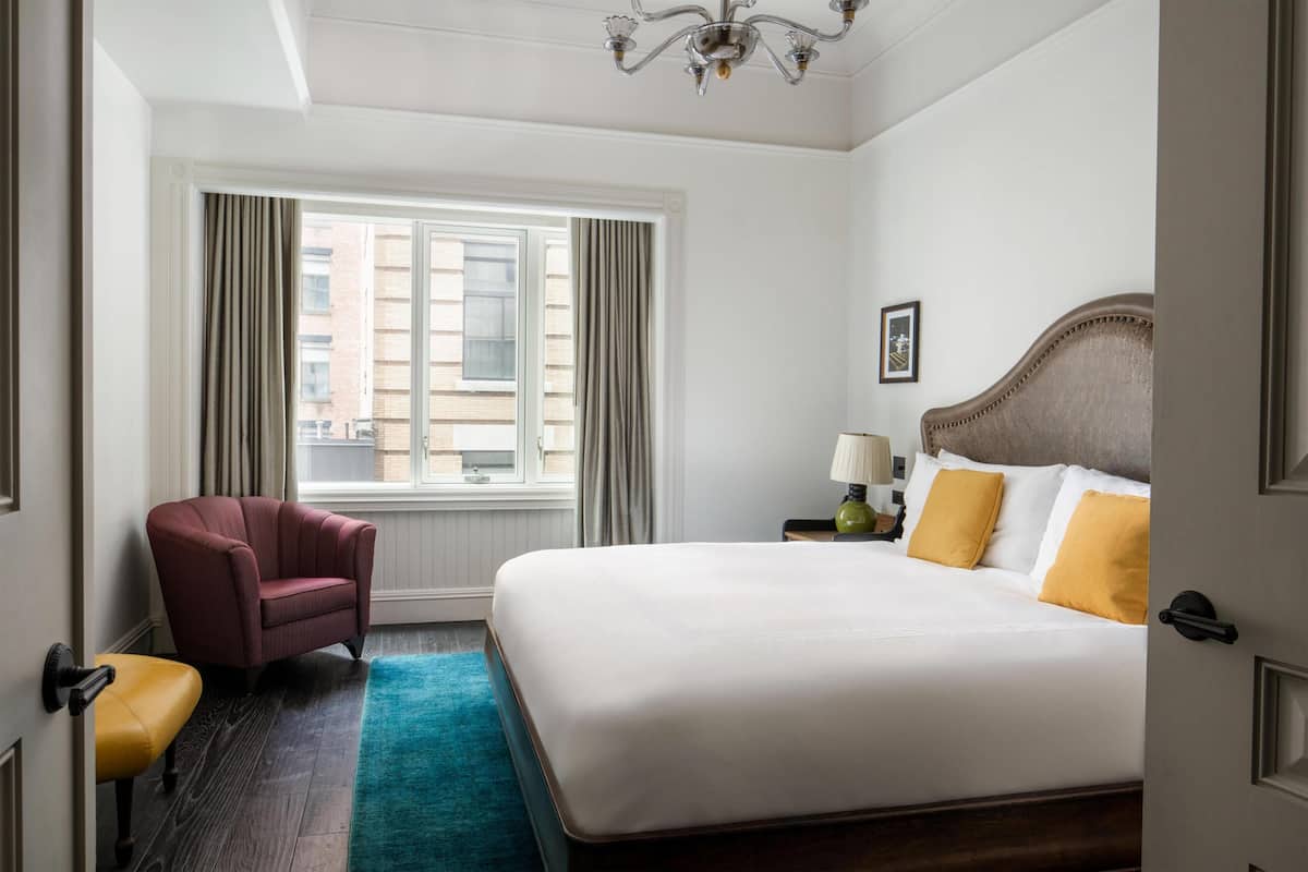 Quarto de hotel com uma cama grande, uma cadeira, uma mesinha e uma janela com cortinas.