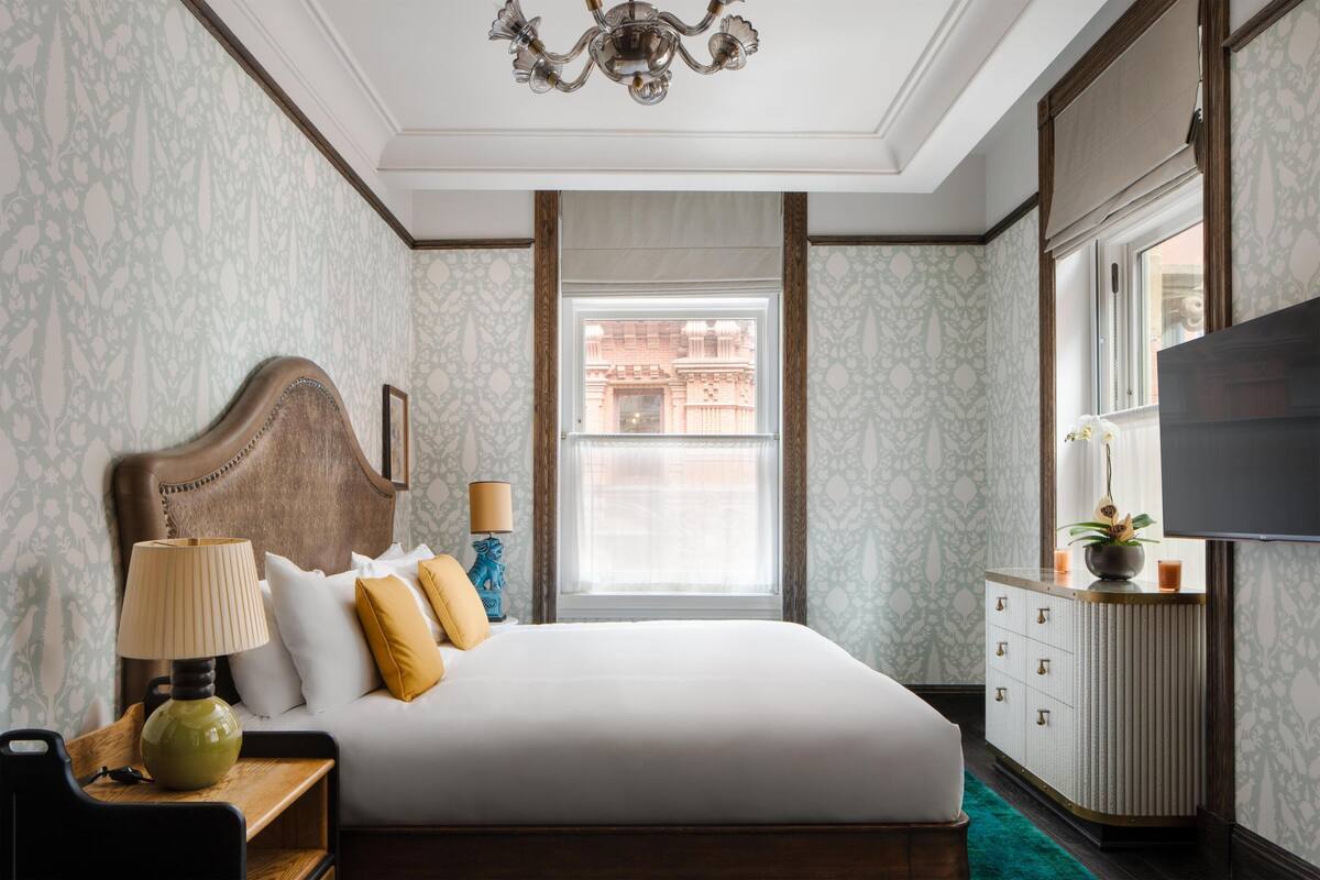 Quarto de hotel com cama, mesa de cabeceira, abajur, cômoda, televisão e janela com vista para edifícios.