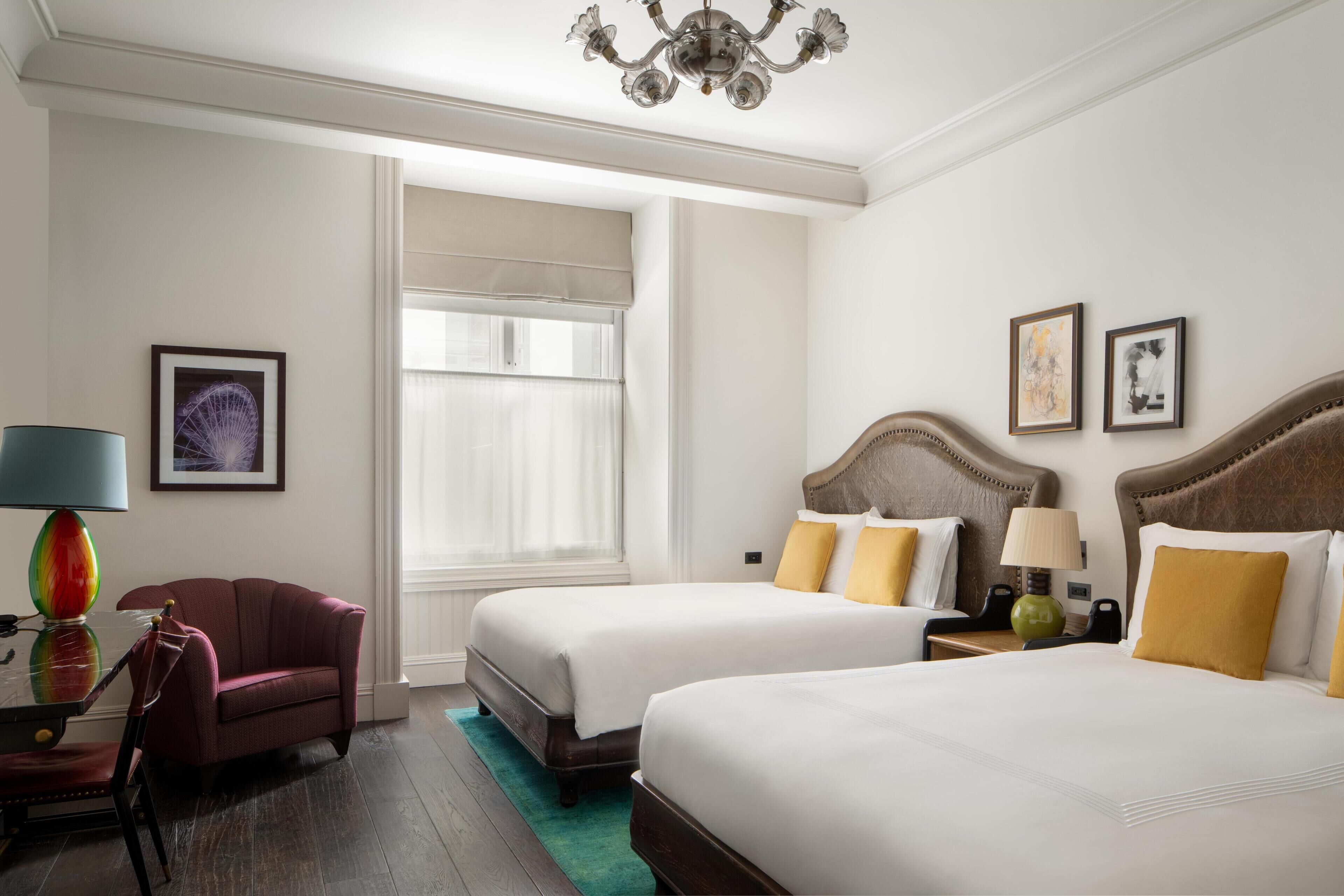 A hotel room with two beds, a desk, a chair, a lamp, and artwork on the walls.