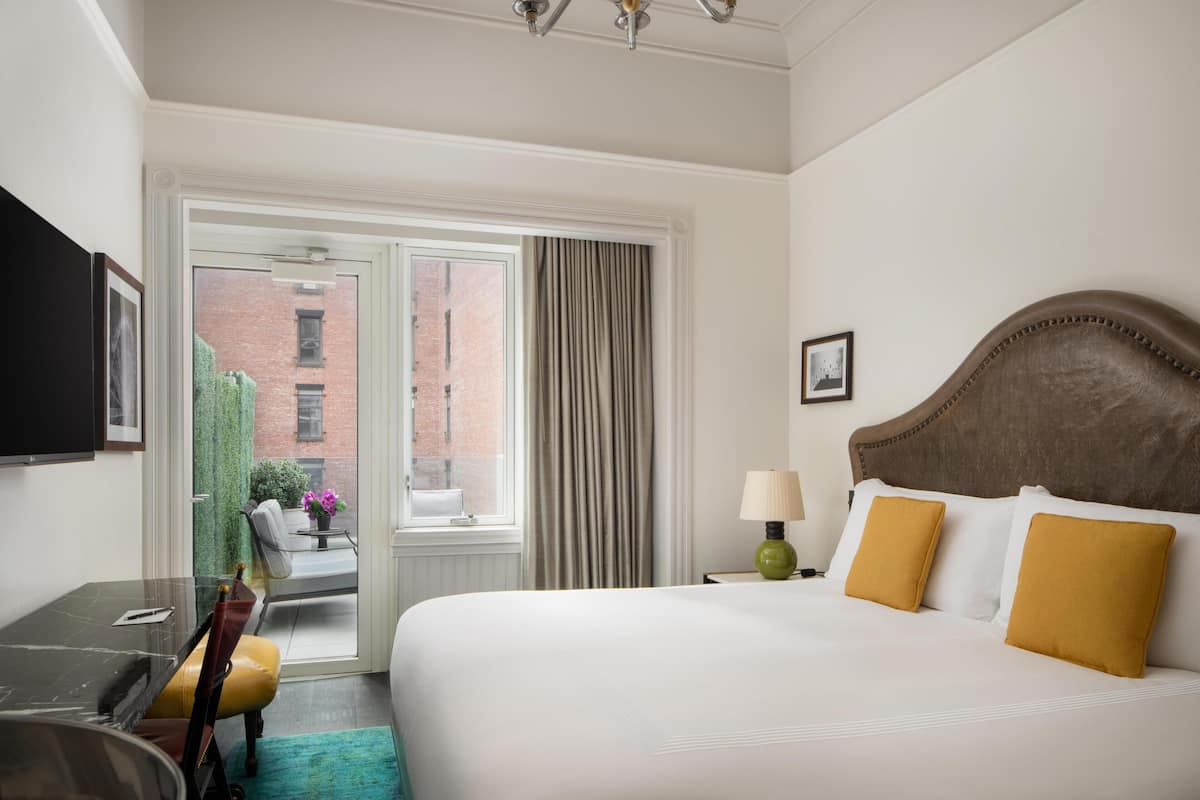 Quarto de hotel com cama, escrivaninha, cadeira, abajur e vista para um edifício de tijolos pela janela.