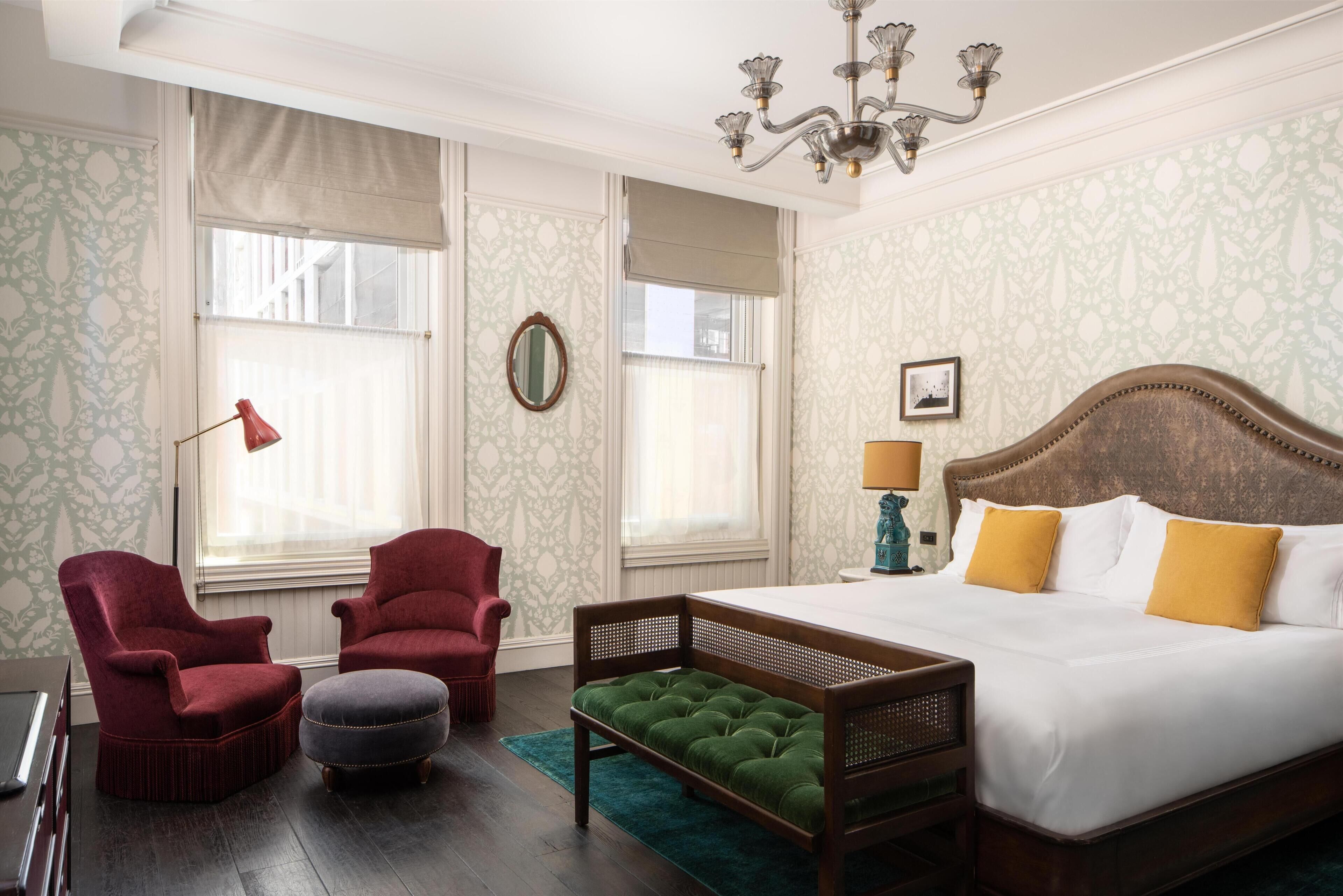 A bedroom with a large bed, two armchairs, a bench, a desk, and a chandelier.